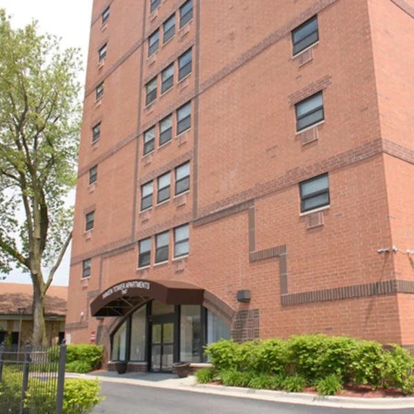 A brick mid-rise apartment building with multiple windows and a covered entrance, surrounded by greenery and a paved walkway.