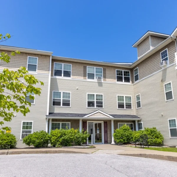Three-story beige apartment building with multiple windows, a covered entrance, and surrounding green shrubs under a clear blue sky.