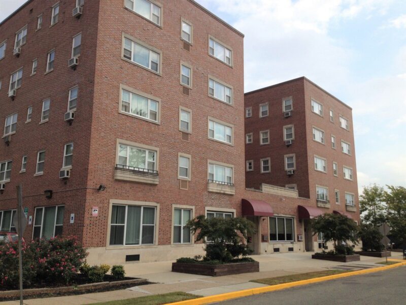 Two four-story brick apartment buildings with large windows, red awnings over the entrances, and landscaping along the sidewalk on a clear day.