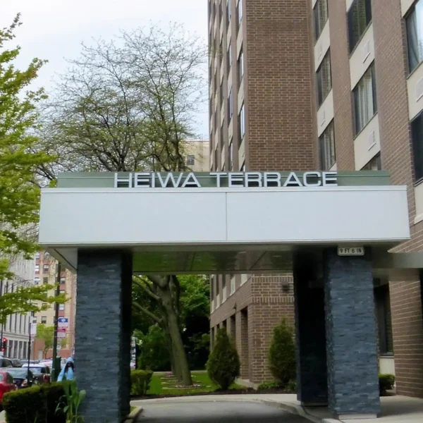 Entrance of a multi-story brick building with a sign reading "Heiwa Terrace" above a covered walkway, surrounded by trees and greenery.
