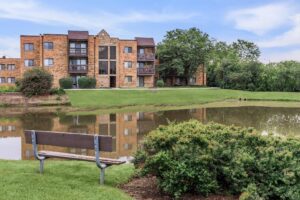 Three-story brick apartment building with balconies overlooks a small pond and grassy area with a bench in the foreground; trees and shrubbery surround the scene.