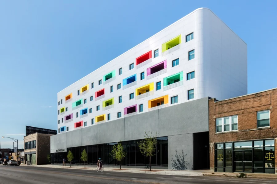 A modern white building with a gray ground floor features colorful balcony recesses in red, blue, green, and yellow; people walk on the sidewalk in front.