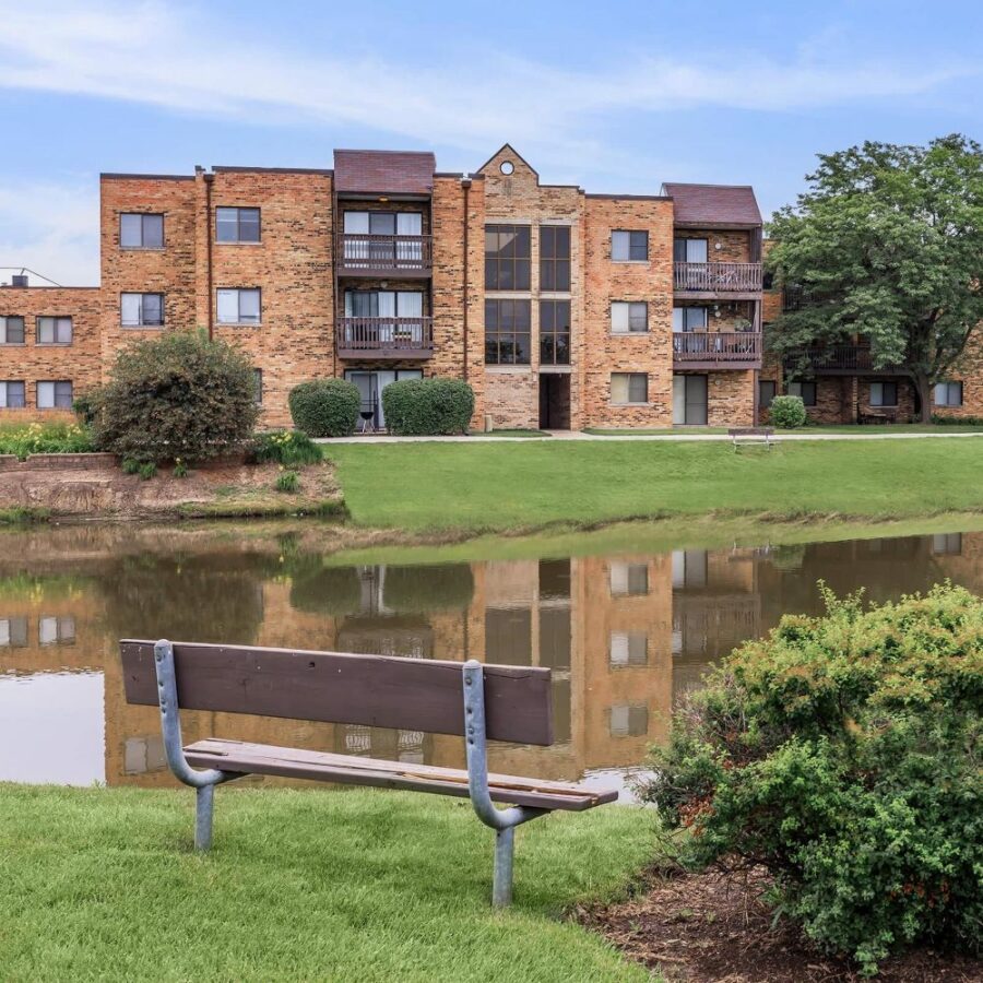 A three-story brick apartment building is reflected in a pond, with a bench and bushes in the foreground and a clear sky above.