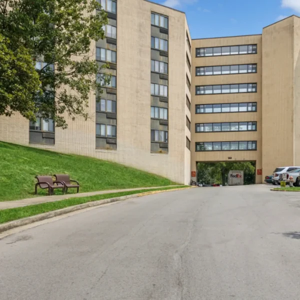 A beige apartment building with six stories, a driveway entrance, green grass, benches, parked cars, and a FedEx truck near the entrance under a blue sky.