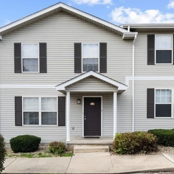 Two-story house with gray vinyl siding, black shutters, a black front door, small covered entryway, and minimal landscaping under a partly cloudy sky.