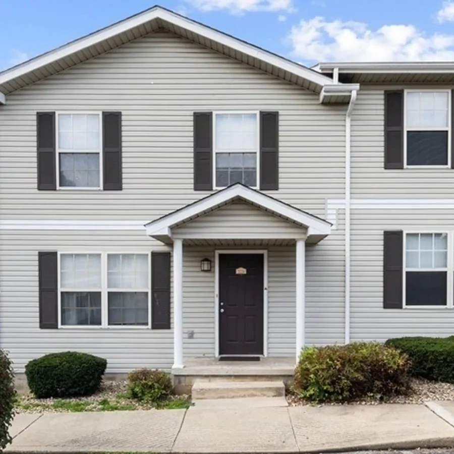 Two-story house with gray vinyl siding, black shutters, a black front door, small covered entryway, and minimal landscaping under a partly cloudy sky.