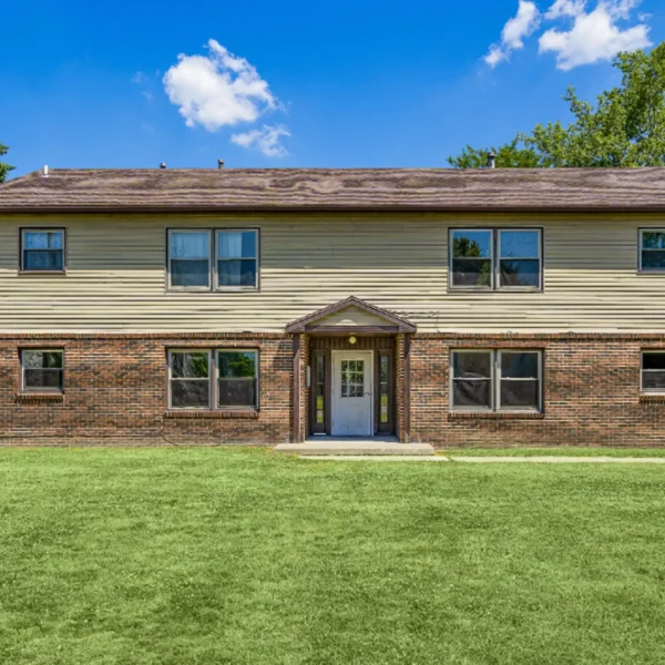 A two-story brick and siding apartment building with four visible windows, a central entrance, and a grassy lawn in front under a blue sky.