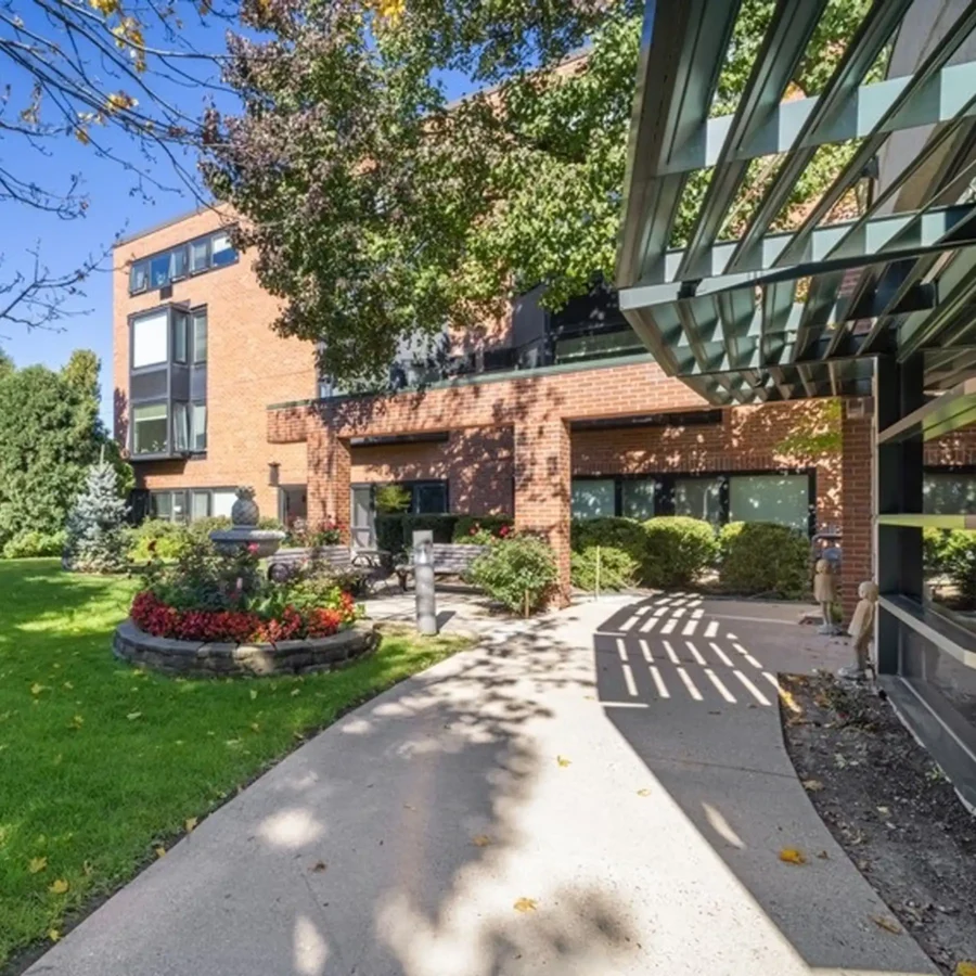 A curved concrete pathway leads to a brick building, bordered by green grass, landscaped plants, and trees under a clear sky.