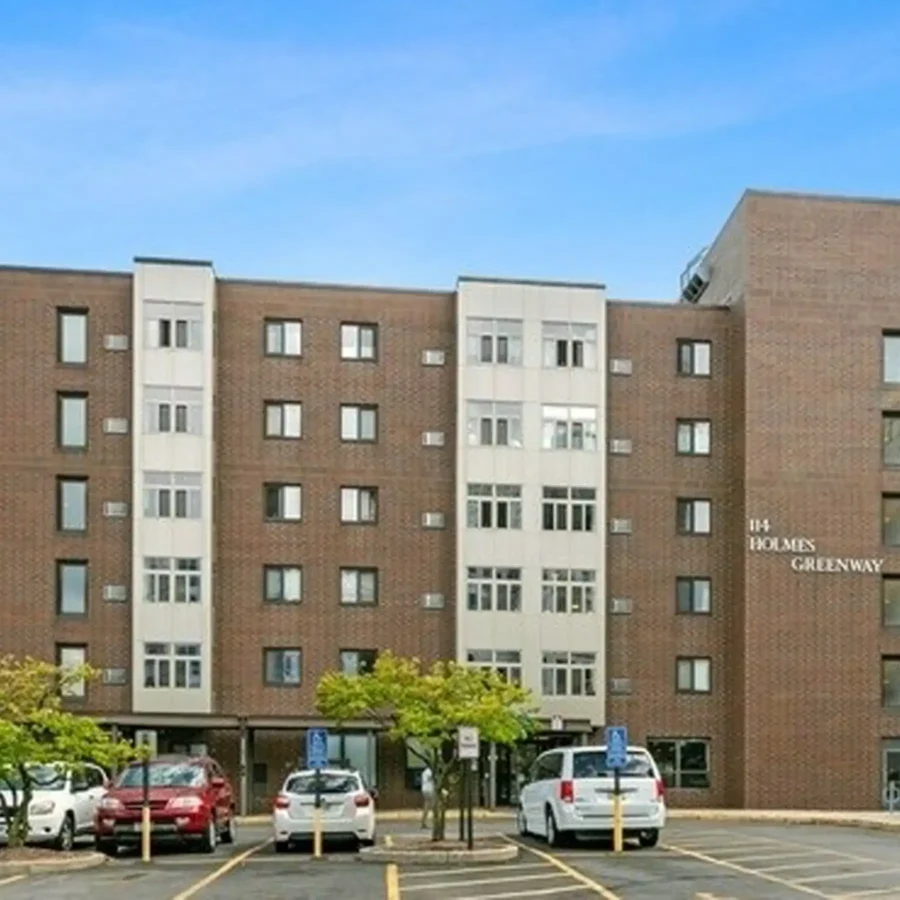A five-story brick apartment building with multiple windows and a sign reading "114 Holmes Greenway," with cars parked in front and trees along the sidewalk.