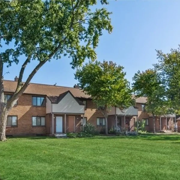 A row of two-story brick apartment buildings with white doors, small front porches, and a well-maintained grassy lawn, bordered by trees under a clear blue sky.