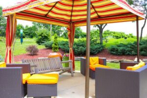 Outdoor seating area under a striped canopy with wicker chairs featuring orange cushions and a wooden bench swing, surrounded by greenery.