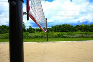 A close-up of a volleyball net set up on a sand court with green trees and a cloudy sky in the background.