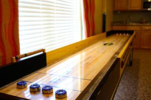 A shuffleboard table with pucks positioned at one end, near a window with blinds and red curtains in a room with a kitchen area.