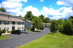 Apartment buildings with beige siding and parked cars along a paved driveway, surrounded by green trees and grass under a partly cloudy sky.
