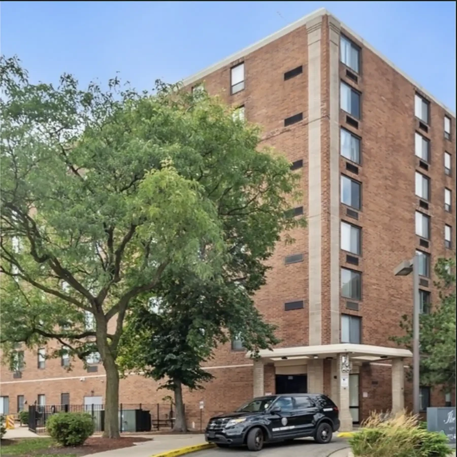 A multi-story brick apartment building with large windows, an entrance canopy, a parked black SUV, and a leafy tree in front.