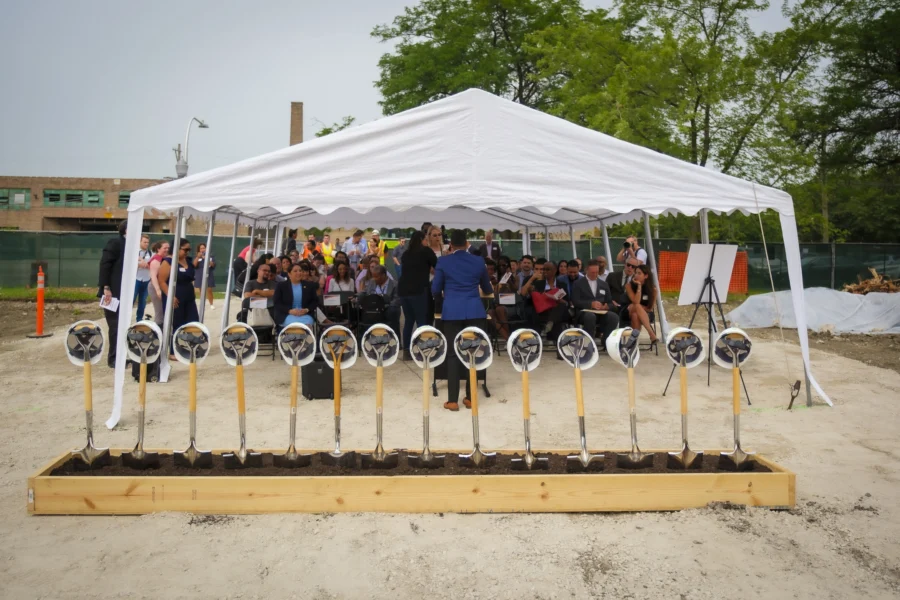 A white tent shelters a seated crowd at an outdoor groundbreaking ceremony, with a row of shovels stuck in dirt in front of the tent.