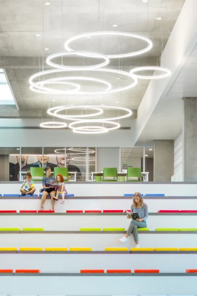 Four people sit on wide, white tiered steps with colorful accents in a modern, bright space; circular light fixtures hang from the ceiling above.