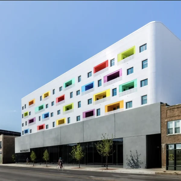 Modern white building with colorful balcony accents, large windows, and a gray ground floor; people are walking and biking along the street in front.
