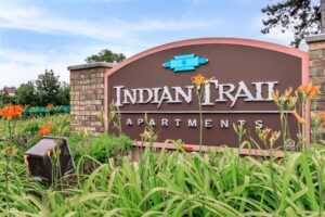 Brown and pink sign reading "Indian Trail Apartments" stands among green plants and orange flowers, with a brick column and trees in the background.