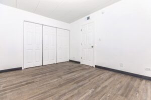 Empty room with white walls, wood-style flooring, a closed white door, and two sets of closed double-door closets.