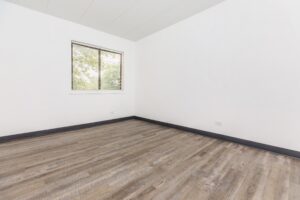 Empty room with white walls, wood-style flooring, a window with blinds, and a black baseboard. Natural light enters through the window.