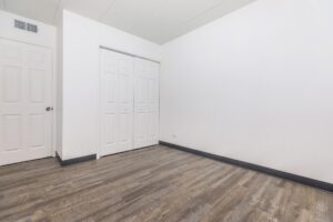 Empty room with white walls, closed white closet doors, a closed white entry door, and wood-style flooring.
