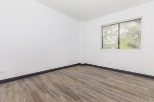 Empty room with white walls, a window with blinds, and wood laminate flooring. Natural light enters through the window, showing an outdoor view with trees.