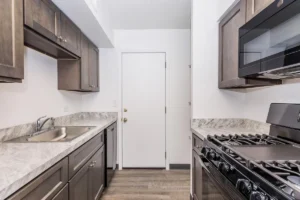 A modern galley kitchen with dark cabinets, marble-look countertops, a stainless steel gas stove, built-in microwave, sink, and a white door at the end.