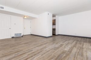 Empty room with light-colored walls, wood-style flooring, two closed doors, and a partial view of a kitchen through an open doorway.