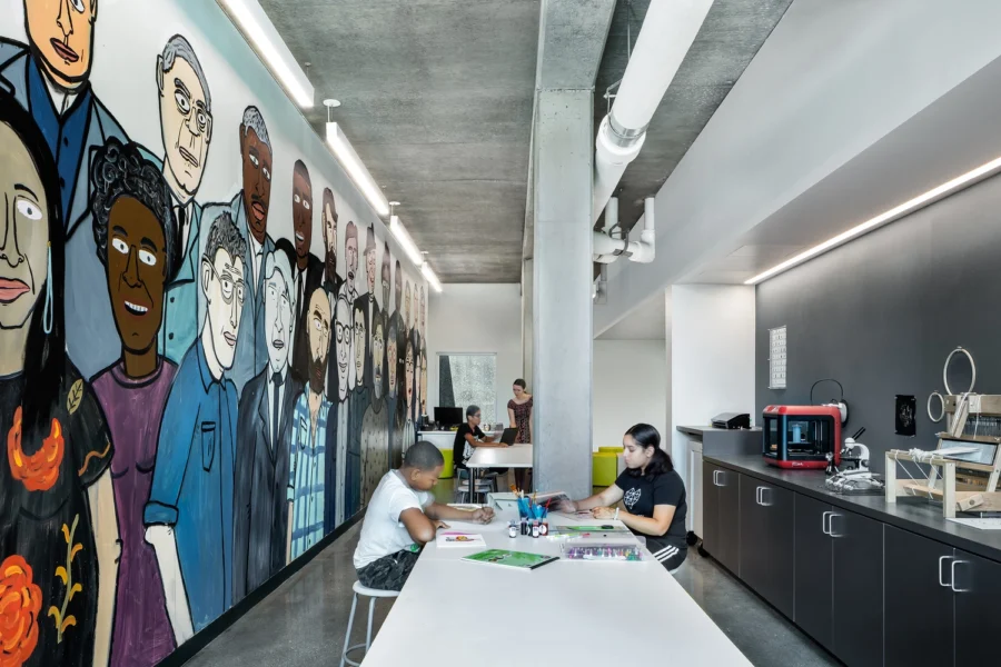 Two people work at a table in a modern room with a mural of diverse faces on the wall; a 3D printer sits on a counter nearby.
