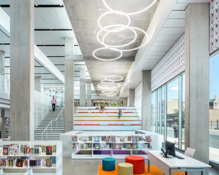 Modern library interior with tall windows, circular ceiling lights, stepped seating area with colorful cushions, bookshelves, and a computer workstation.