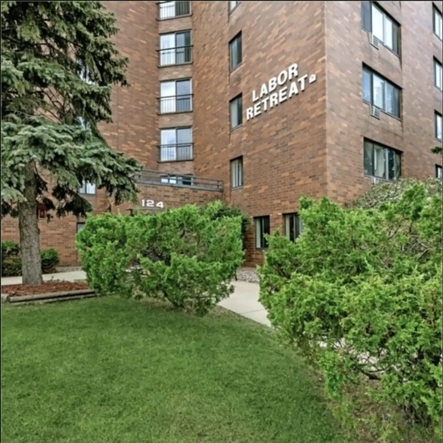 A brick apartment building with "LABOR RETREAT" written on the exterior, surrounded by green bushes and a tree near the entrance.