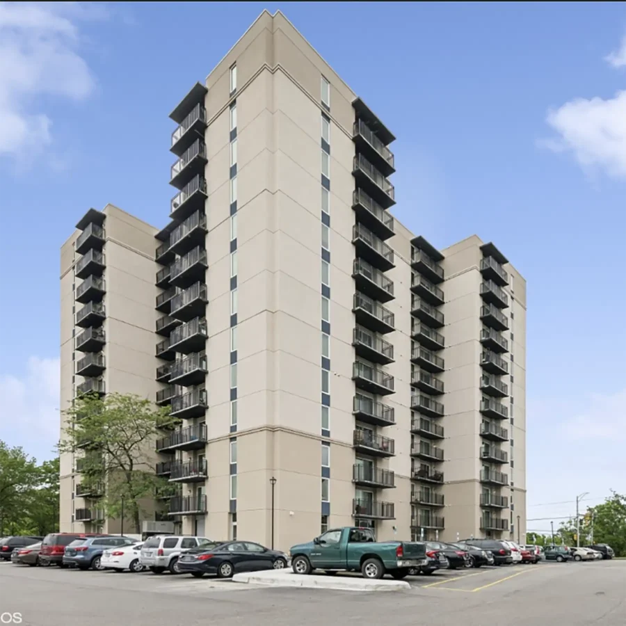 A tall beige apartment building with multiple balconies and rows of parked cars in a lot under a clear, blue sky.
