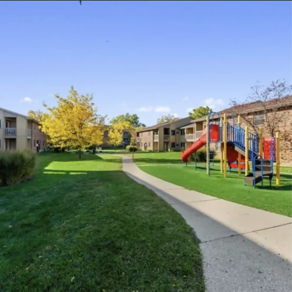 A paved walkway runs between two apartment buildings with green lawns, trees, and a playground with slides and climbing equipment on the right.