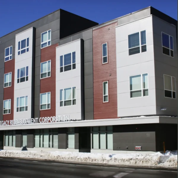 A modern four-story apartment building with large windows, a mix of gray, white, and red panels, and snow piled along the sidewalk.