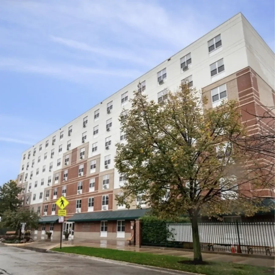 A six-story brick and white building with a green awning, trees in front, and a pedestrian crossing sign on a cloudy day.