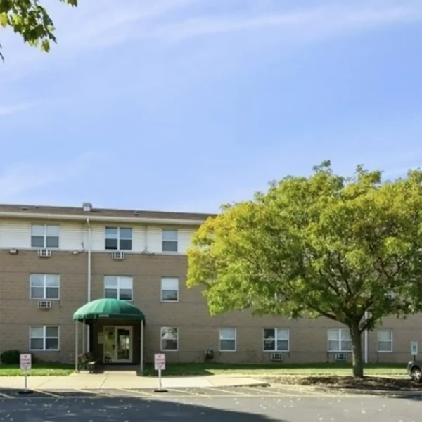 A three-story brick apartment building with a green awning over the entrance, a large tree nearby, and a parking lot in front.