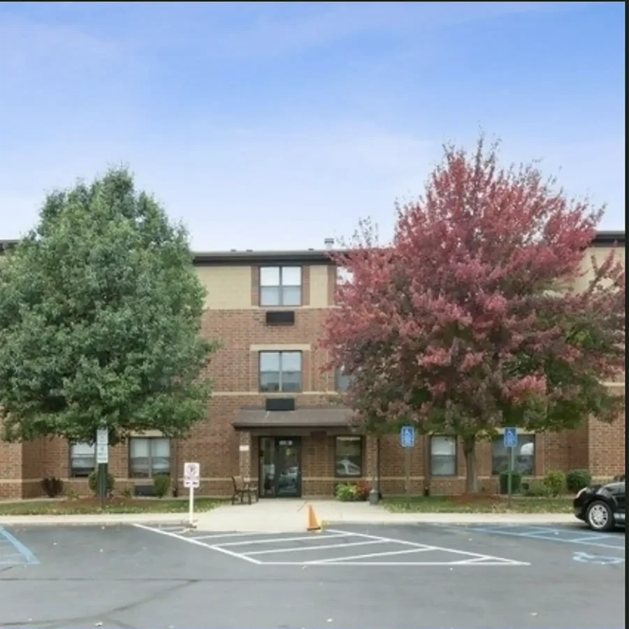 A three-story brick building with a central entrance, flanked by two trees—one green and one red—in front, and an empty parking lot with handicap spaces.