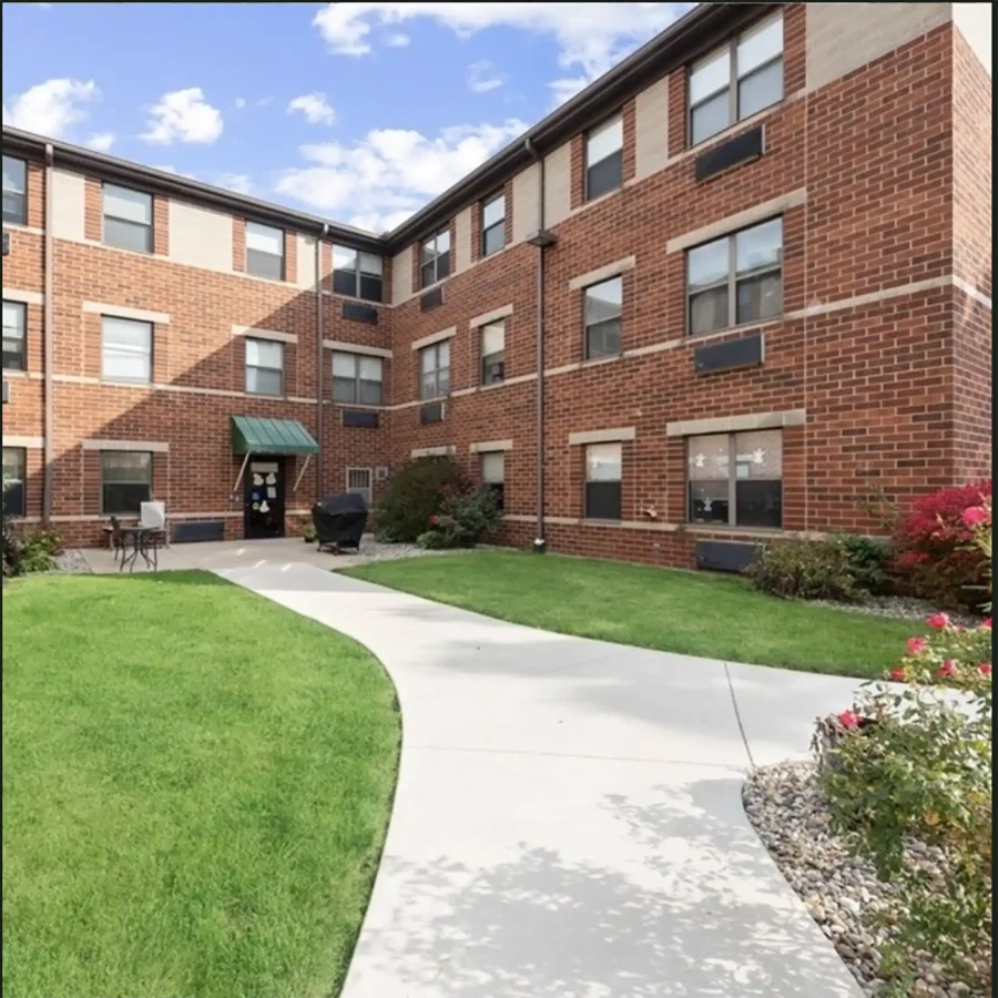 A paved walkway leads to the entrance of a three-story red brick apartment building with multiple windows and landscaped greenery.