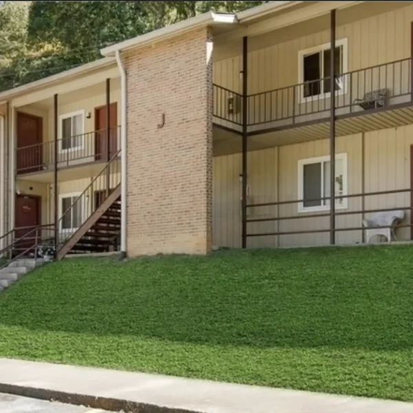 Two-story apartment building with brick and siding exterior, outdoor stairway, and a grassy slope in front. Balcony railings and several windows and doors are visible.