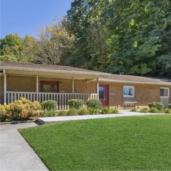 Single-story brick building with a covered front porch, red doors, and landscaped grass lawn, set against a backdrop of tall trees.