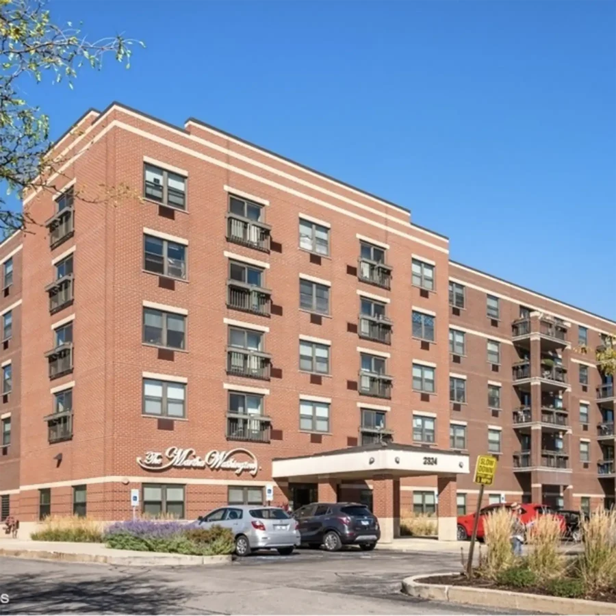 A four-story red brick apartment building with balconies, parked cars in front, and a clear blue sky overhead.