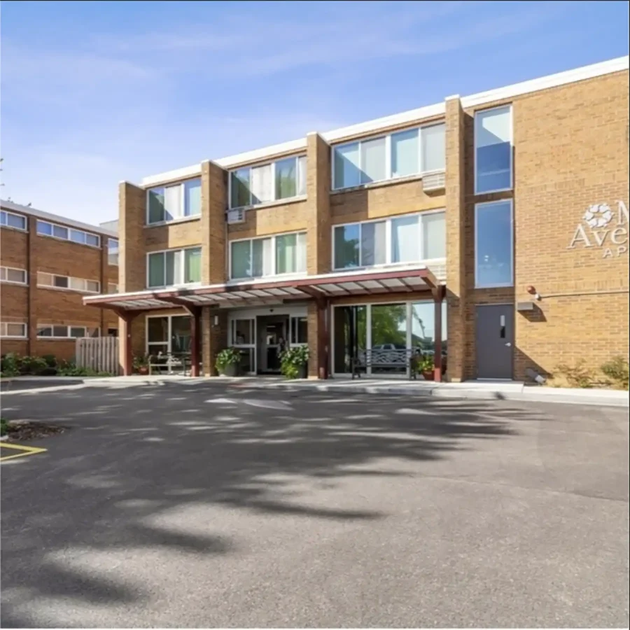 A three-story brick apartment building with large windows, a covered entrance, potted plants, and a paved driveway in front.