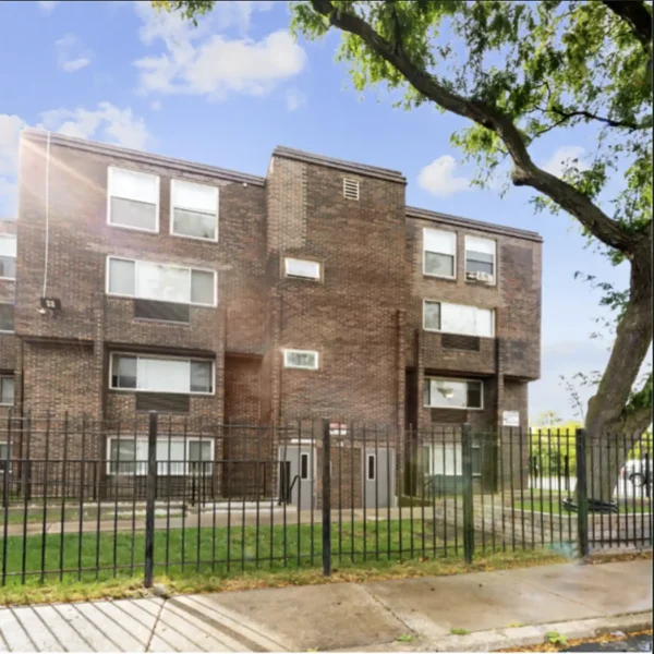 Three-story brick apartment building with large windows, metal fence in front, and a tree on the right side under a partly cloudy sky.