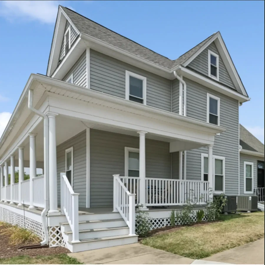 A two-story gray house with white trim and a large wraparound porch, featuring steps leading up and neatly trimmed bushes along the front.