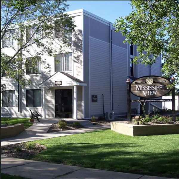 A three-story apartment building with white siding and a sign reading "Mississippi View" stands next to a green lawn and trees.