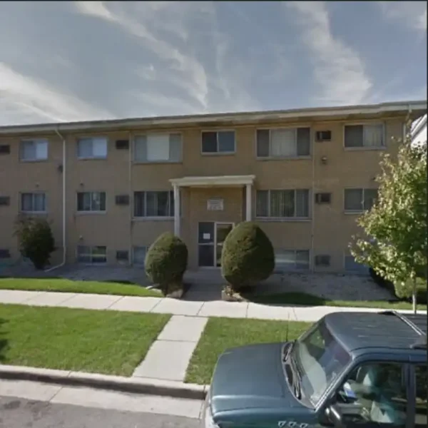 A two-story beige brick apartment building with trimmed bushes in front, a small entrance canopy, and a green vehicle parked at the curb.
