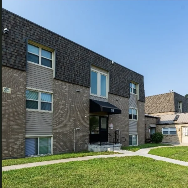A three-story apartment building with brown brick exterior, shingle siding on the top floor, multiple windows, and a black entrance canopy, surrounded by grass and sidewalks.