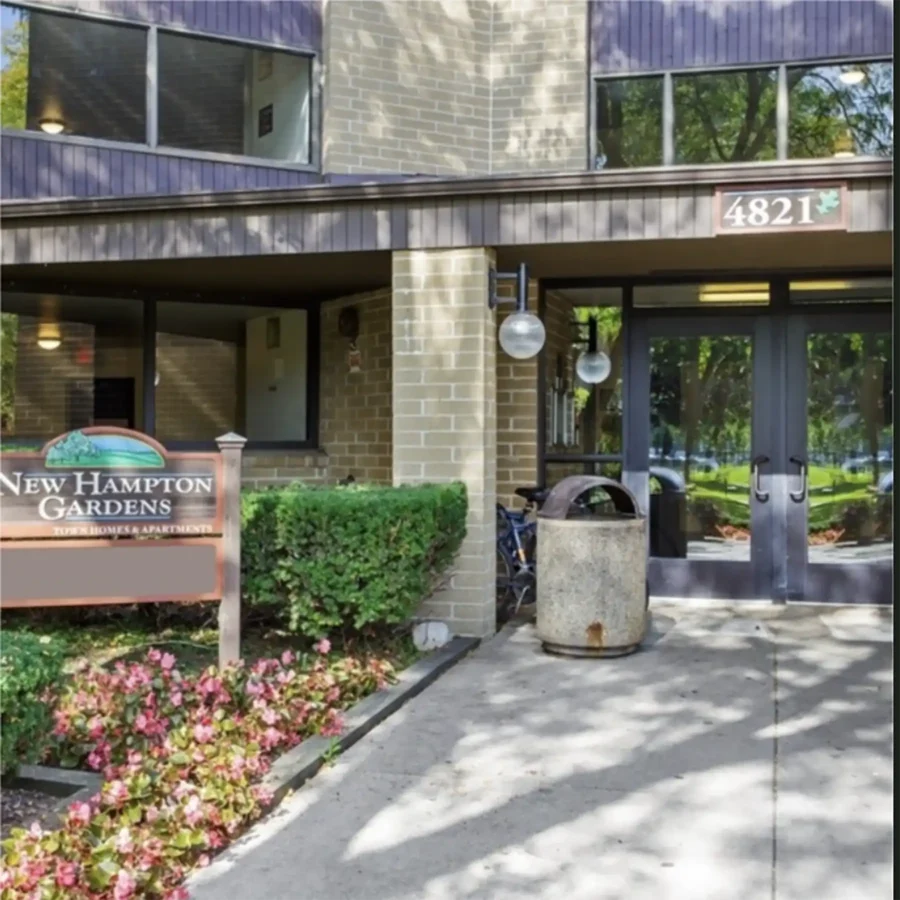 Entrance of an apartment building with glass doors, address number 4821, a "New Hampton Gardens" sign, a trash can, plants, and flowers along the walkway.