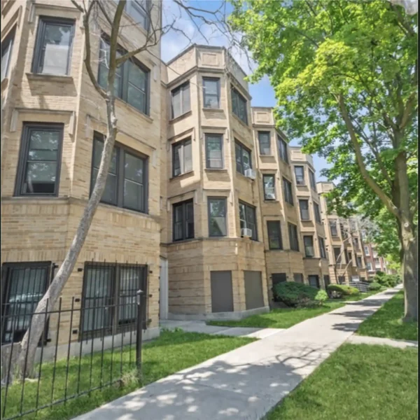 A row of multi-story brick apartment buildings with bay windows, black window frames, and a tree-lined sidewalk on a sunny day.